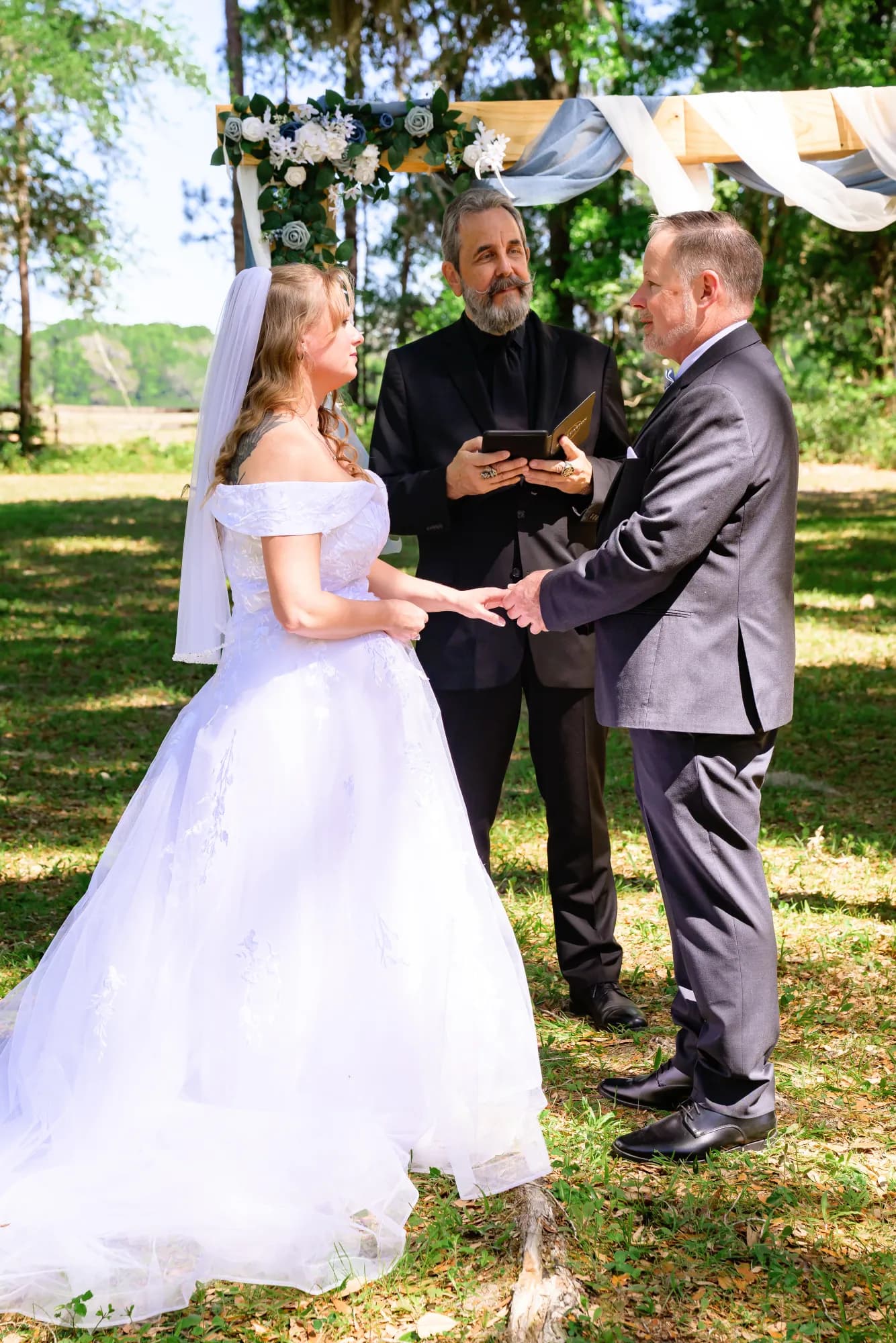A bride in a full skirted gown and veil holding both hands of her groom in a grey suit while an officiant in black reads from a book between them, standing on green grass beneath a timber arch dressed with blue and white flowers and white drapes.