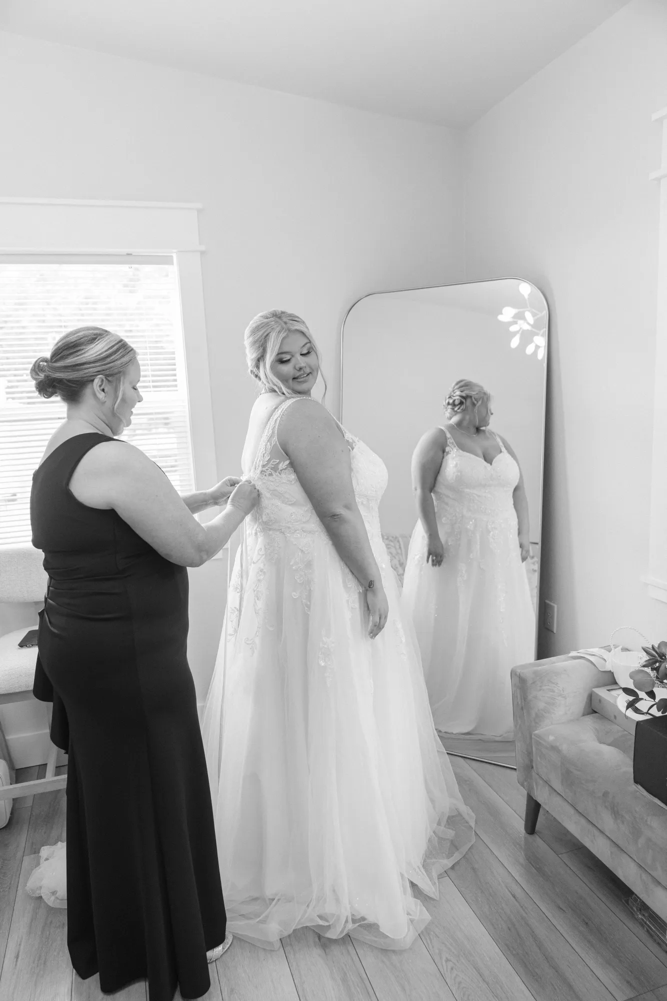 Black and white photograph of a bride looking back over her shoulder while her mother fastens the back of her lace gown, the pair standing beside a tall arched floor mirror in the bridal suite that reflects the full length of the dress.