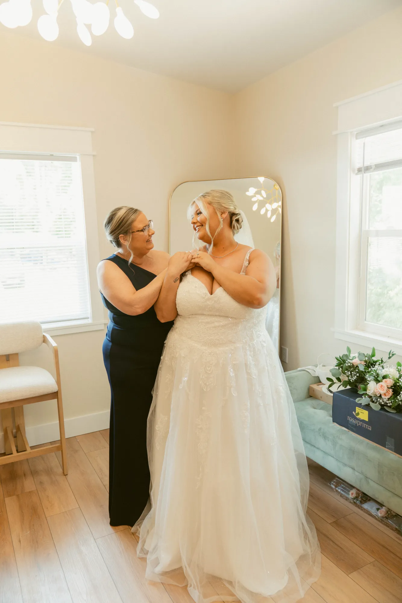 A bride in a lace gown standing in the bridal suite holding her mother's hands, both laughing, in front of a tall arched floor mirror between two bright windows, with pale wood floors, a pale green settee and a bouquet resting on it.