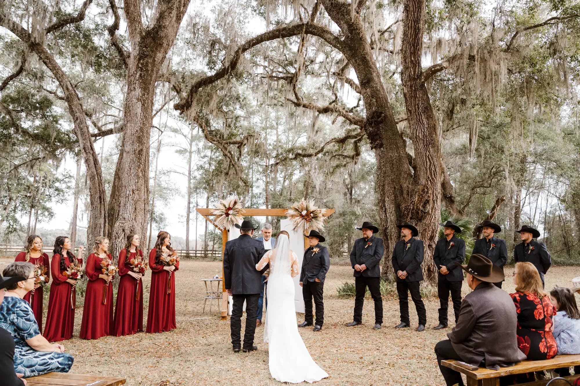 A wedding ceremony in progress beneath two spreading live oaks, the couple facing each other at a timber arbor with attendants in deep red and black standing to either side.