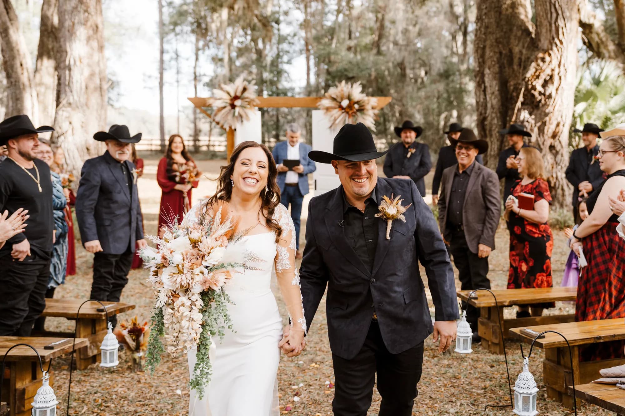 A newly married couple walking hand in hand back up the grass aisle, laughing, guests applauding from wooden benches on either side.