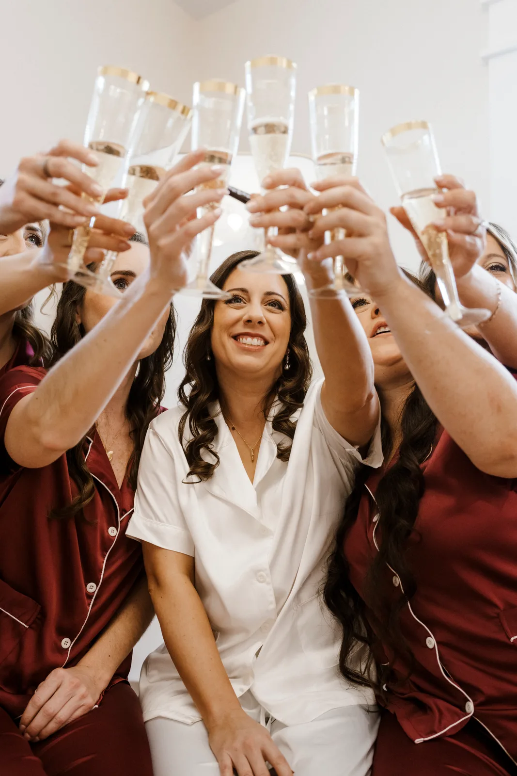 A bride in a white satin pajama set smiling in the middle of her bridesmaids in burgundy pajamas, everyone raising gold-rimmed champagne flutes together toward the camera.
