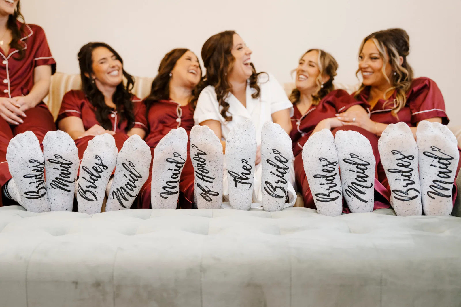A bride and her four bridesmaids laughing on a sofa in matching burgundy and white pajamas, their feet up in front, with socks reading Bridesmaid, Maid of Honor and The Bride.