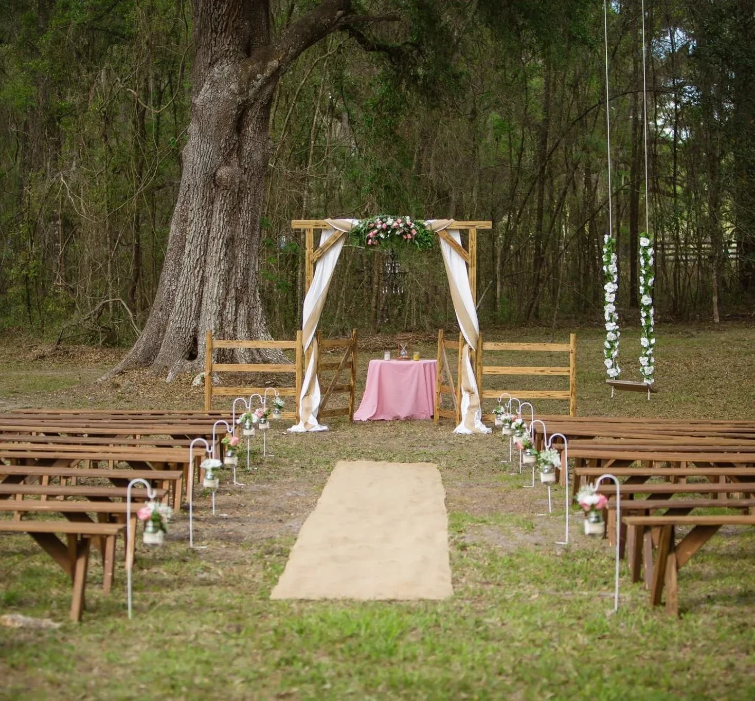 A floral ceremony arch draped in white fabric under a spreading oak, with wooden benches either side of an aisle runner