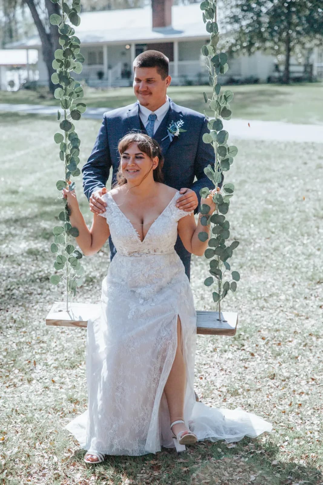 A bride seated on a rope swing hung from an oak, her groom standing behind her with his hands on her shoulders, the farmhouse beyond.