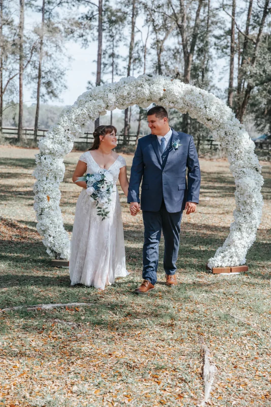 A bride and groom walking hand in hand through a tall circular arch covered in white flowers, the tree line behind them.