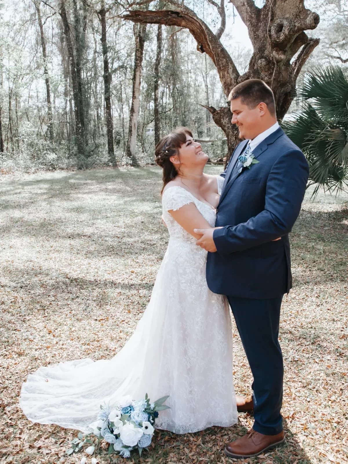 A bride and groom holding each other under the moss hung oaks, her bouquet of white roses resting at her side, fallen leaves across the grass.