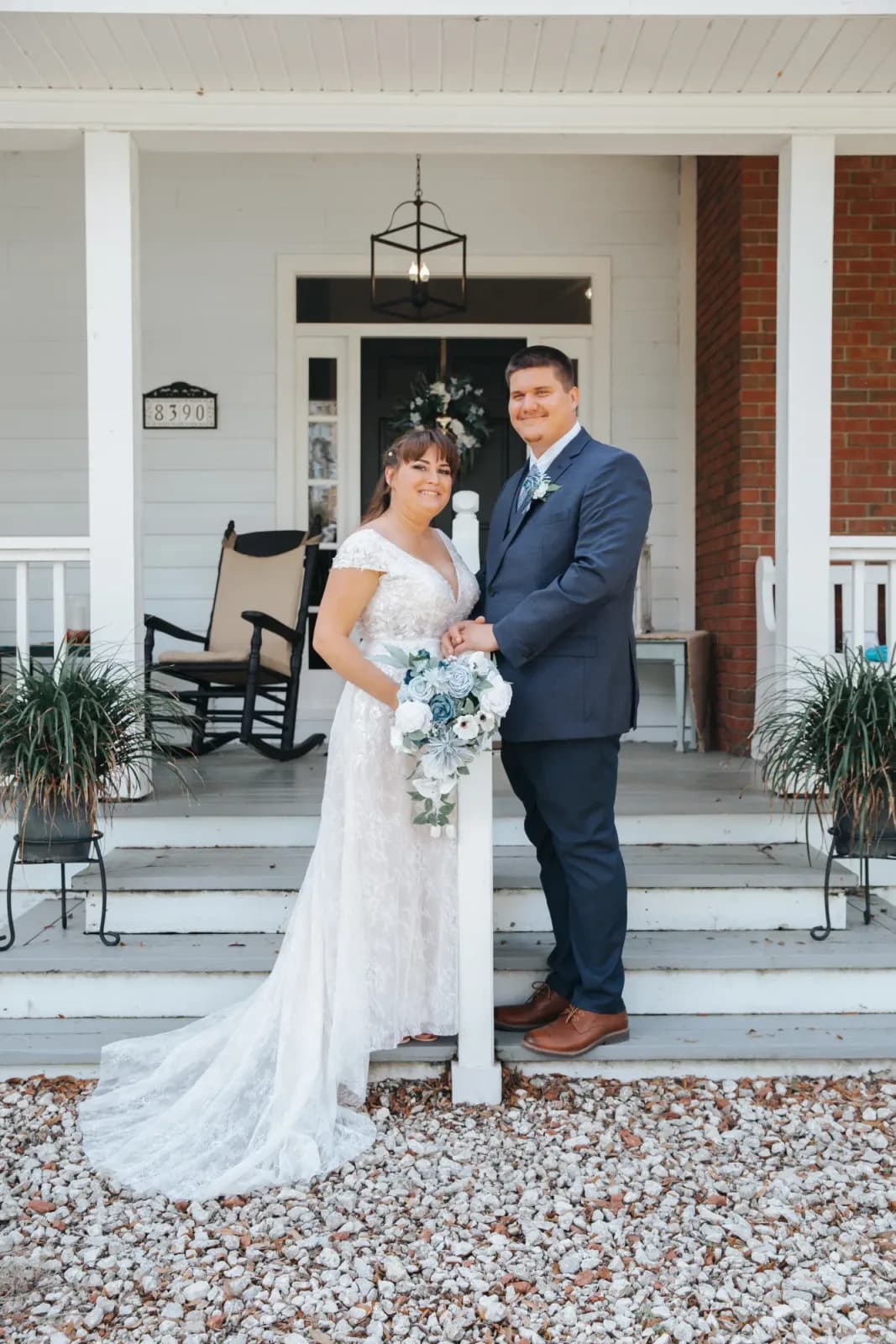 A bride and groom standing together at the foot of the farmhouse steps, rocking chairs and hanging ferns on the porch behind them.