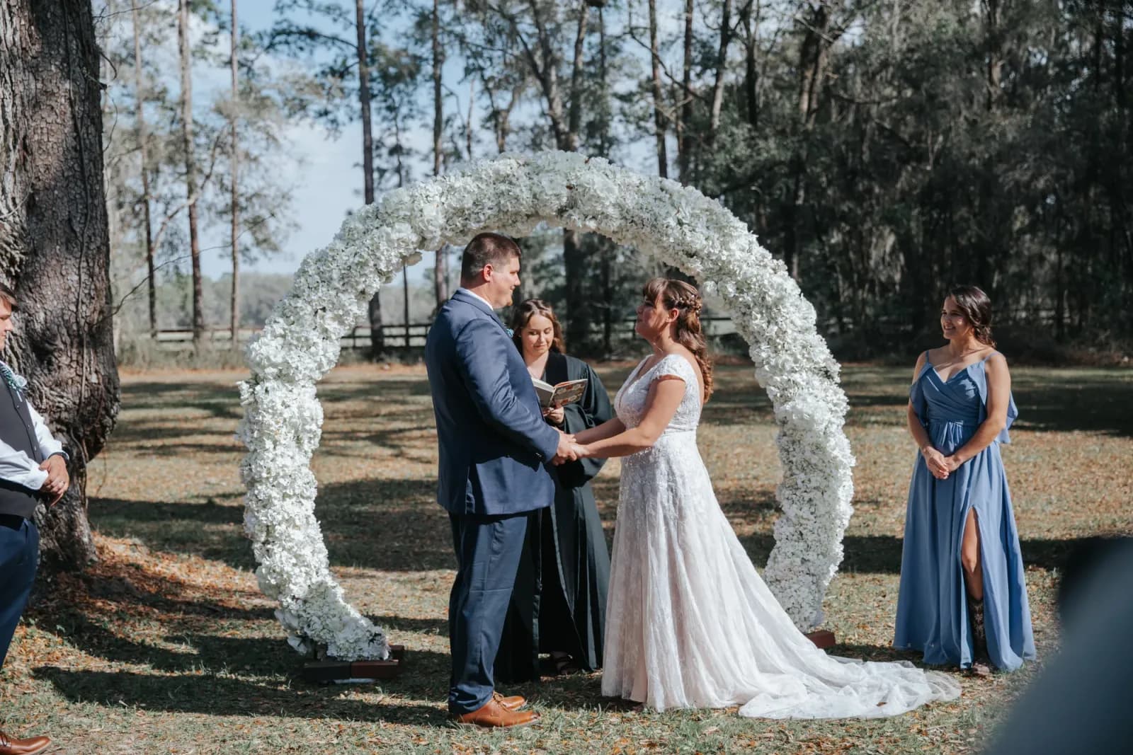 A couple exchanging vows in front of the officiant beneath a circular white floral arch, a bridesmaid in dusty blue standing to the side.
