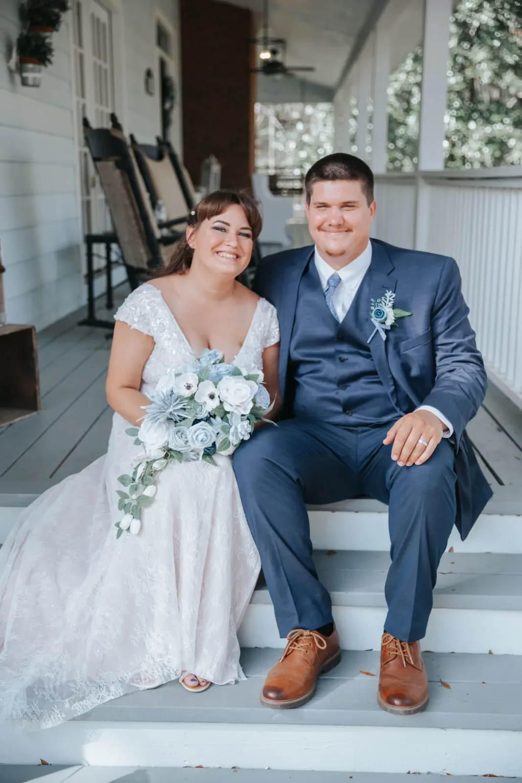 A bride and groom sitting side by side on the farmhouse steps, she in lace with a blue and white bouquet, he in a navy suit.