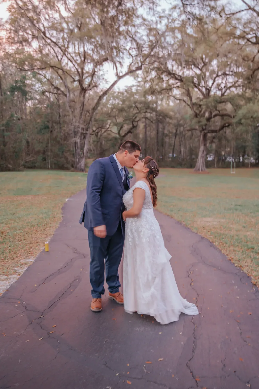 A bride and groom kissing in the middle of the oak lined lane at golden hour, her train spread out on the paving behind her.