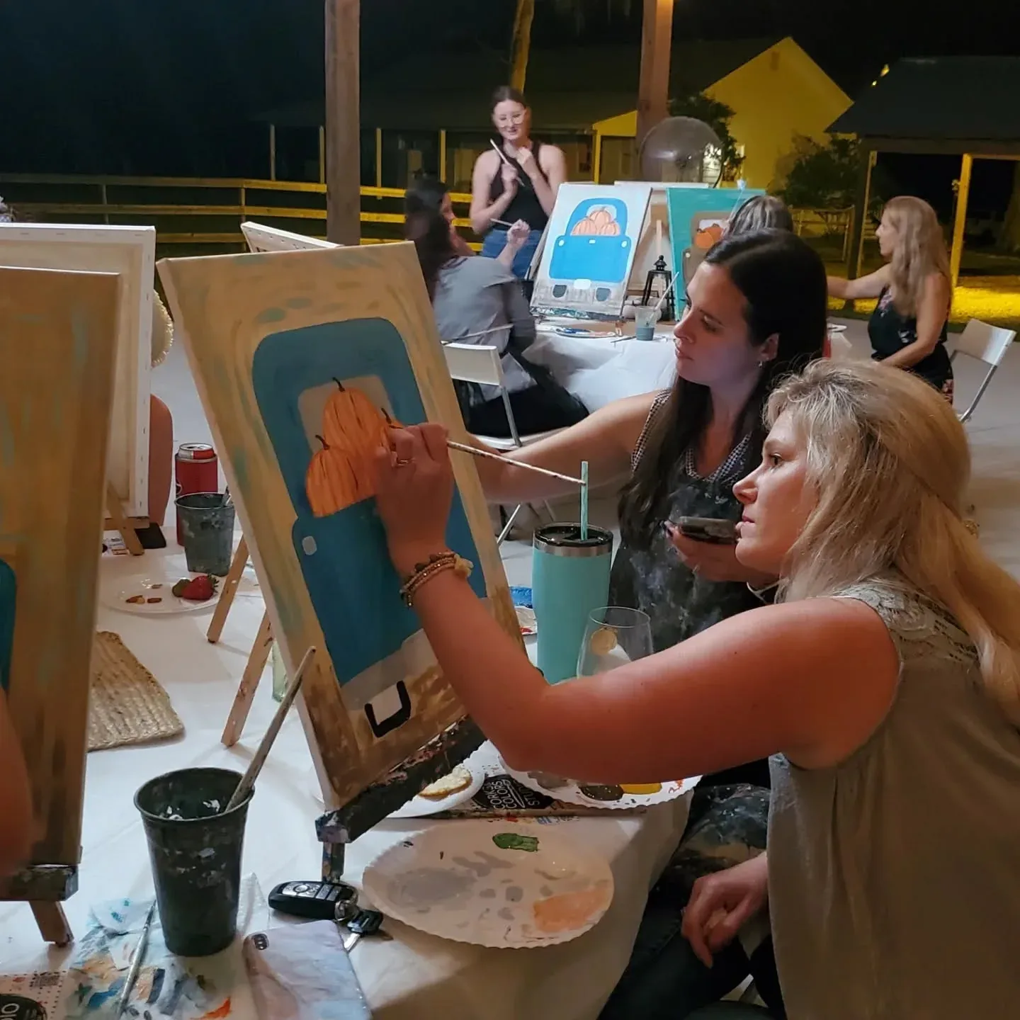 Two women painting at easels during a class, palettes and brushes on the table