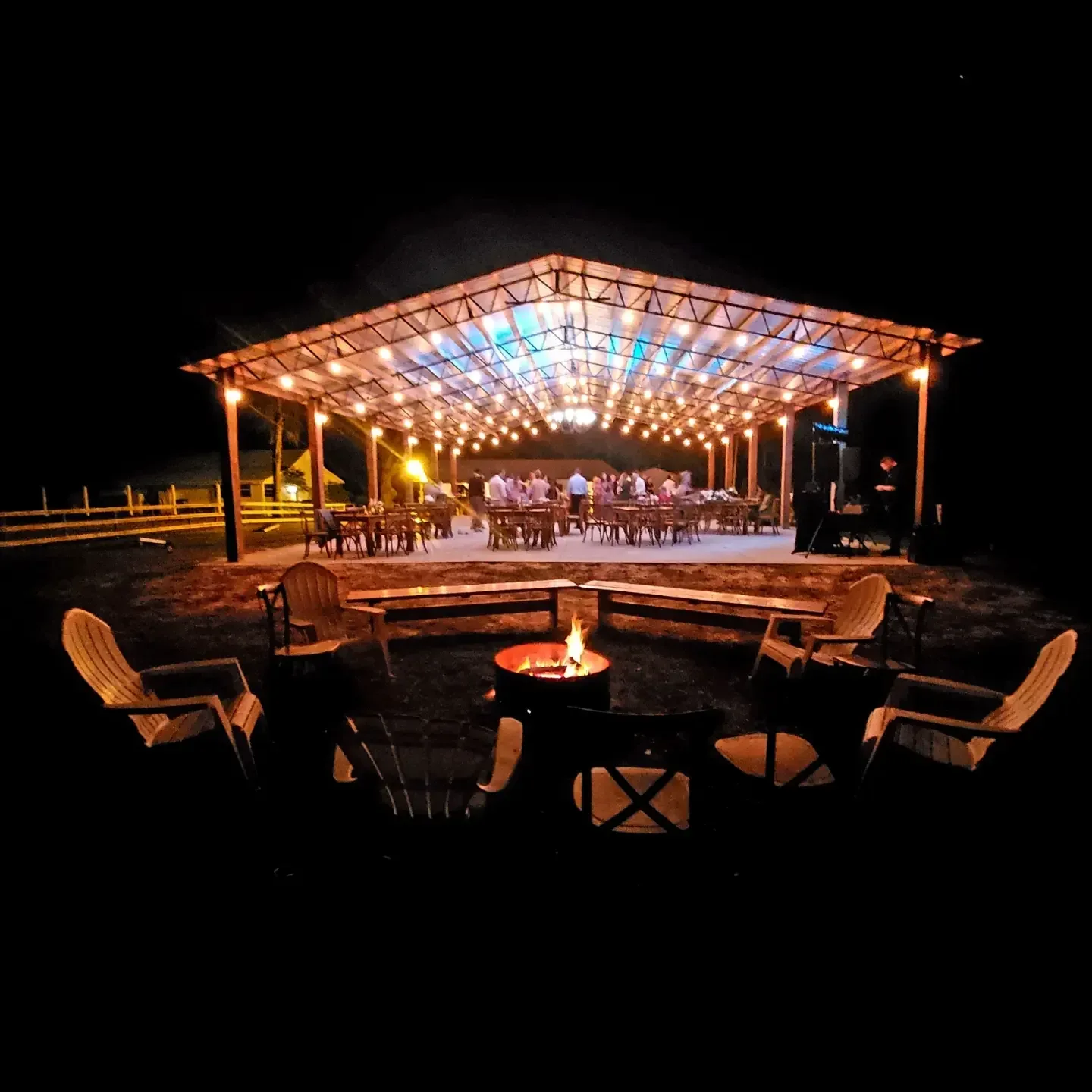 The pavilion lit up at night, its roof beams glowing above rows of tables