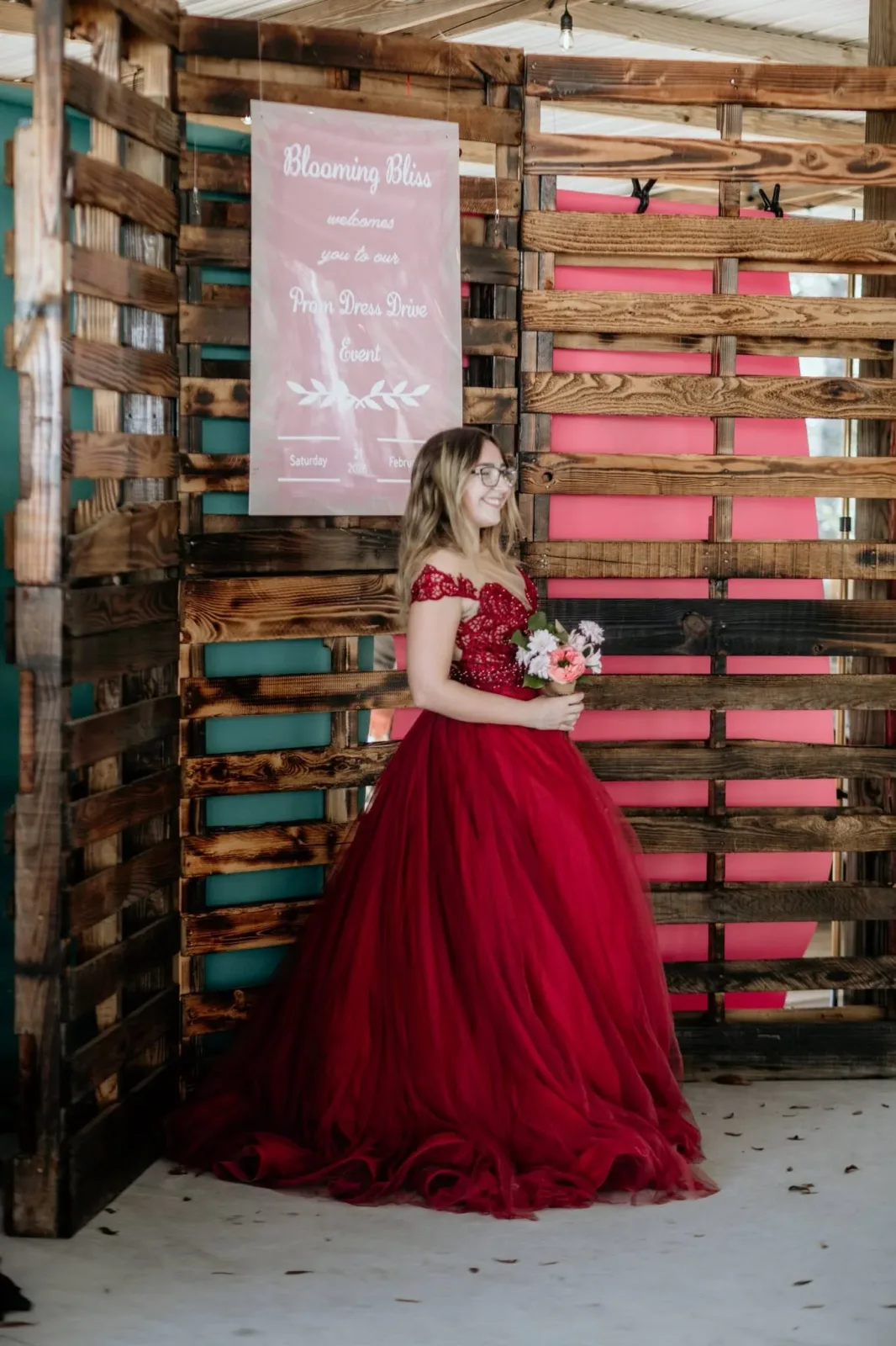 A young woman in a deep red ball gown standing in front of the pallet backdrop at the prom dress drive
