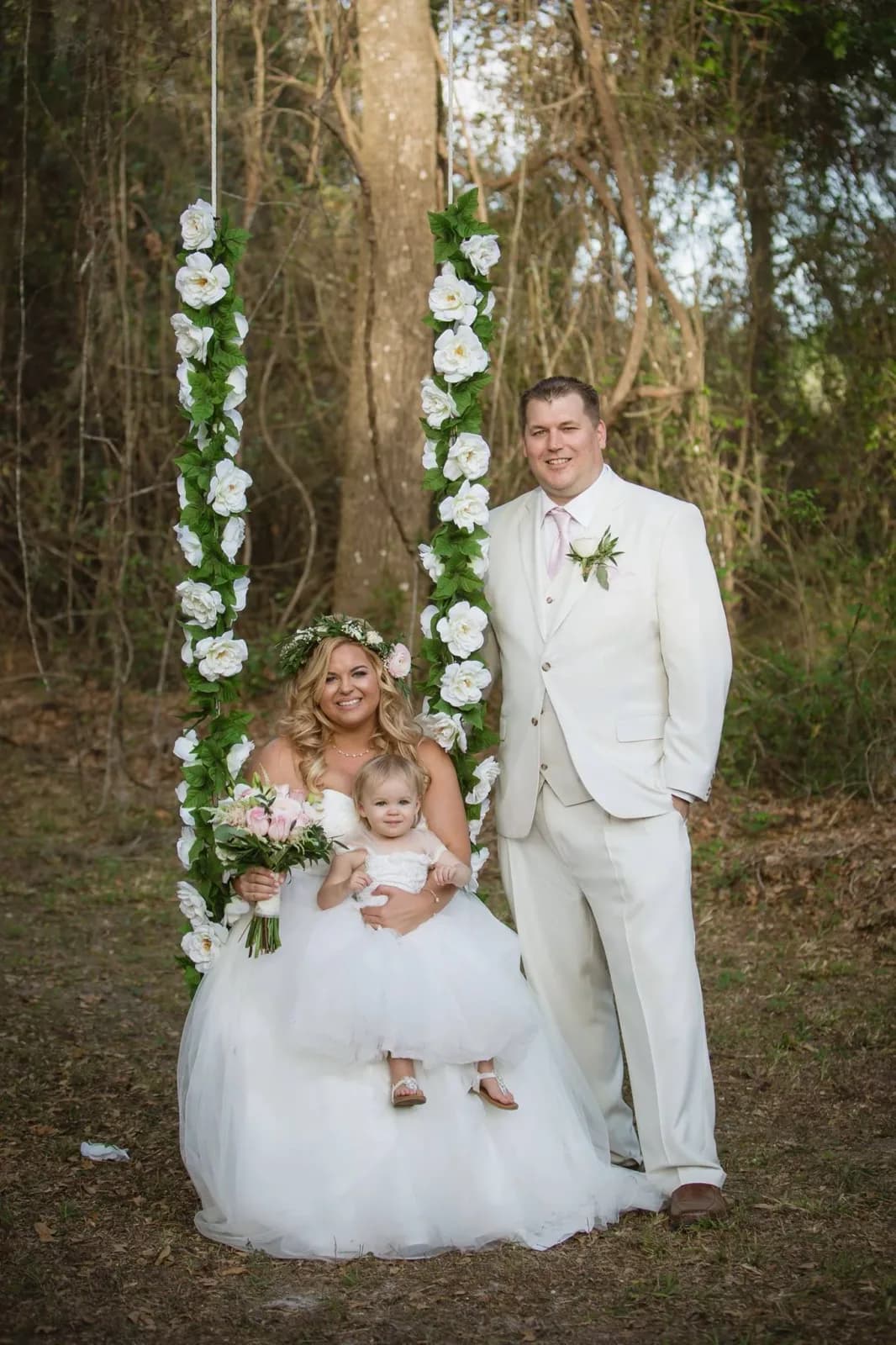 A bride and groom with a small child, framed by a floral column beside the oaks