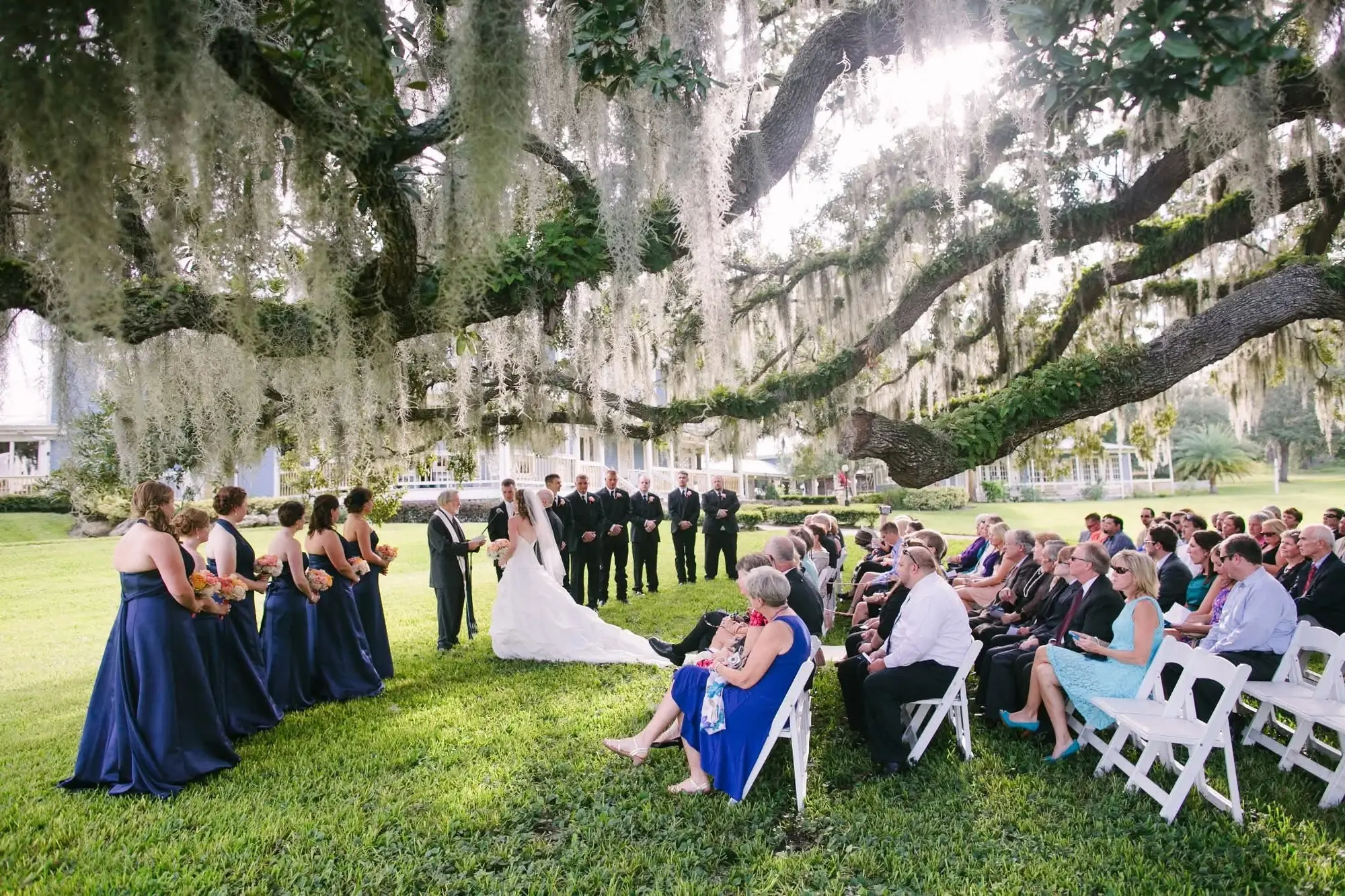 Betsy and Adam's wedding ceremony in 2012, the couple standing before an officiant on the grass beneath a huge moss draped live oak, bridesmaids in navy to one side and guests seated in white chairs, a white farmhouse in the distance.