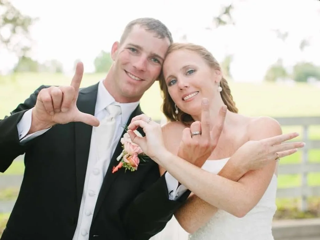 Betsy and Adam on their wedding day, cheek to cheek and smiling at the camera, holding up their hands to make an L shape and showing their new rings, a white fence and open field behind them.