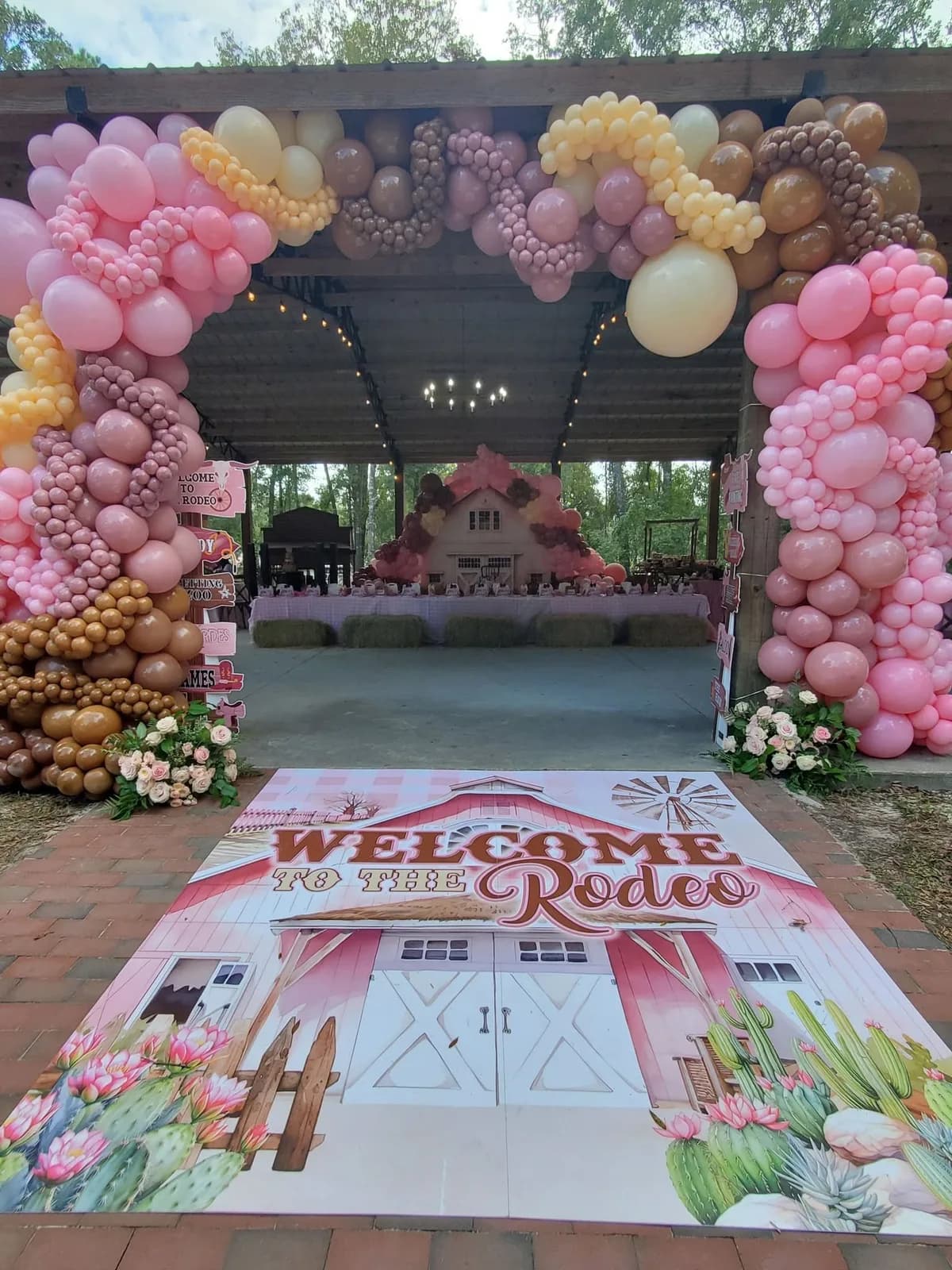 A pink and gold balloon arch framing the entrance to the pavilion, with a floor decal reading Welcome to the Rodeo laid on the brick path in front of it and a pink barn backdrop at the far end.