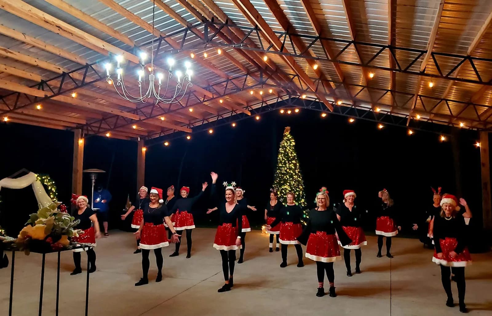 A line of dancers in black tops and red skirts mid-routine on the pavilion floor, a lit Christmas tree and strings of warm bulbs behind them.