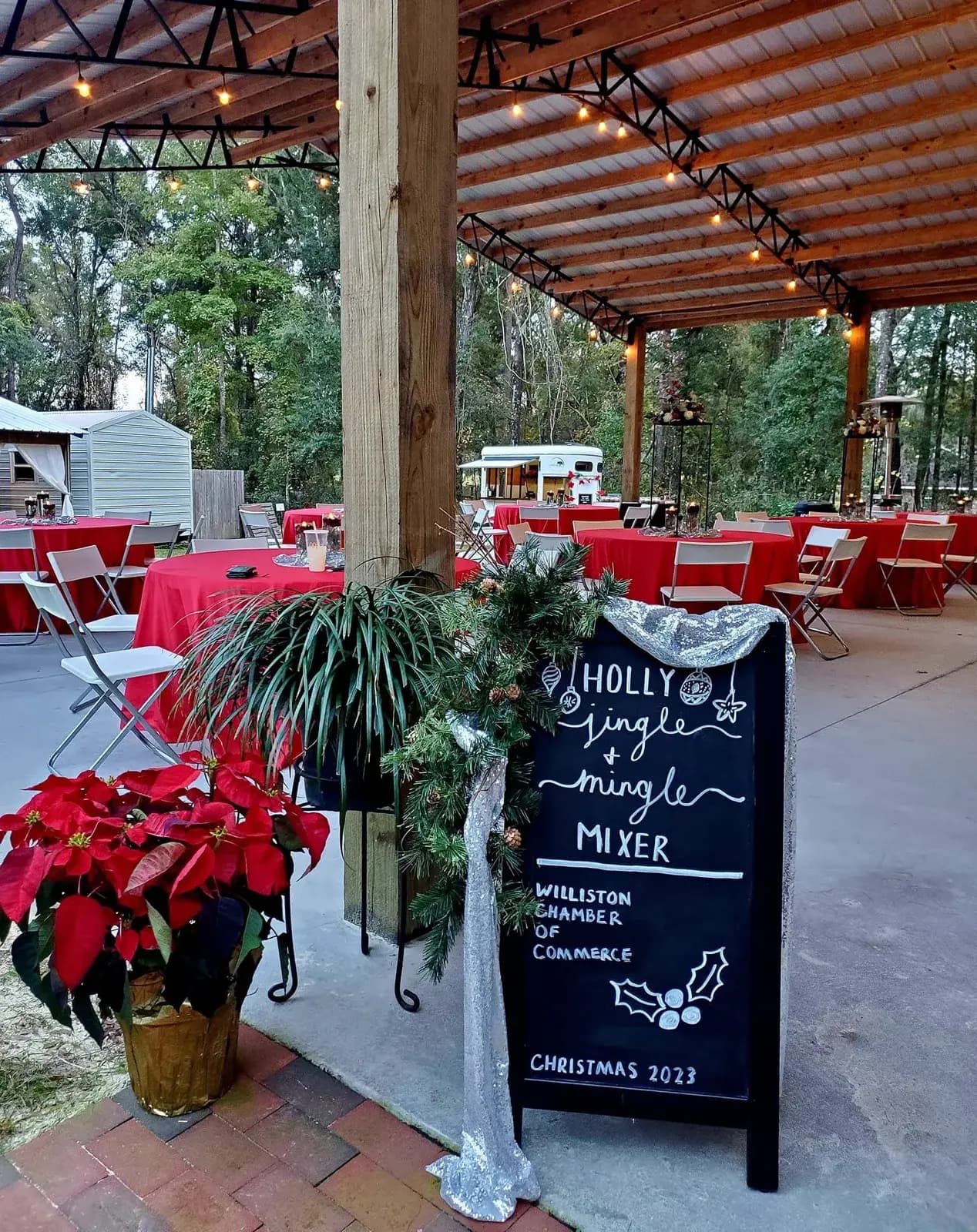 A chalkboard sign reading Holly Jingle and Mingle Mixer, Williston Chamber of Commerce, Christmas 2023, standing beside a bank of red poinsettias, with red-clothed tables set under the pavilion behind it.