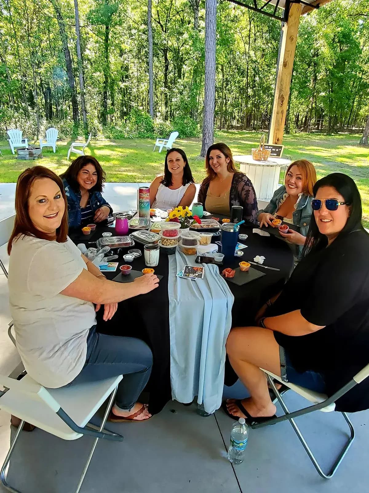 A group of women seated around a table under the pavilion roof in daylight, charcuterie boards and drinks in front of them, mown lawn and woods behind.