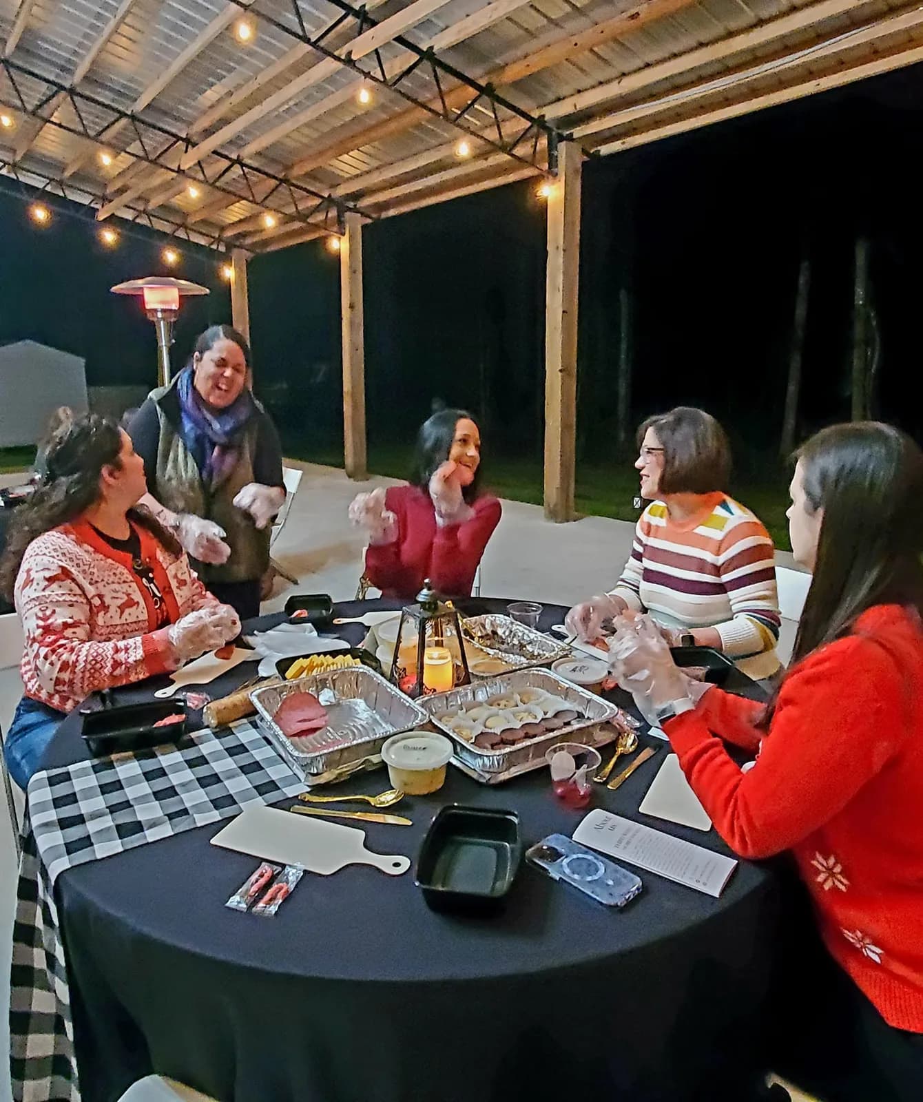 Five women in gloves building charcuterie boards at a black-clothed table at night, trays of meat, cheese and fruit spread between them, string lights and a patio heater behind.