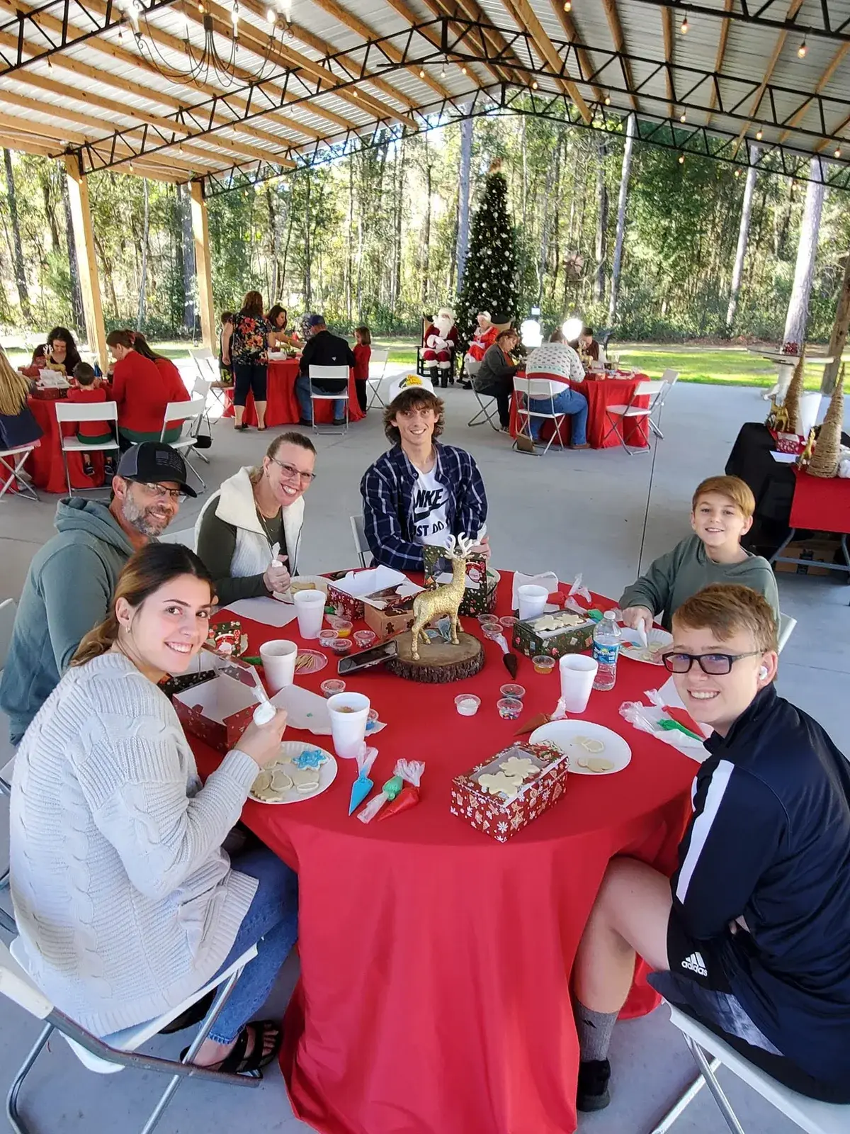 Several families seated at red-clothed round tables across the pavilion floor decorating cookies, a decorated Christmas tree and the open woods at the far end.
