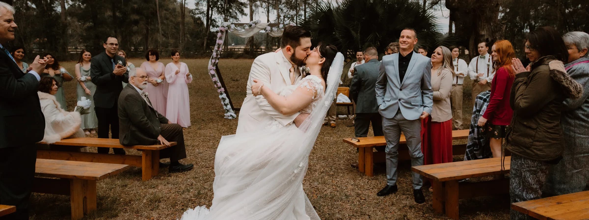 A bride and groom kissing in the middle of the grass aisle at the end of their ceremony, guests standing and applauding from wooden benches on both sides, the flower draped arbor and the treeline behind them.