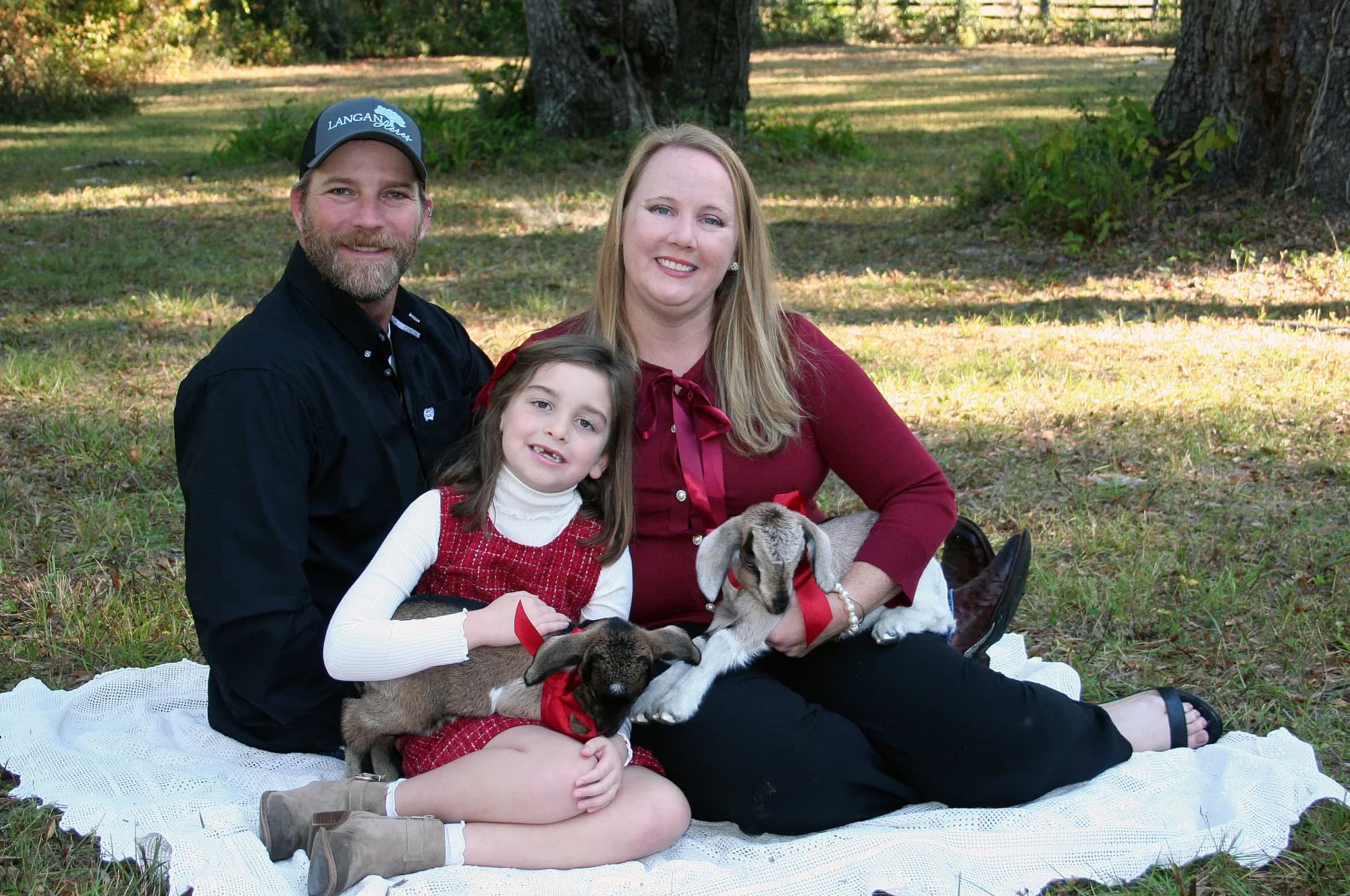Betsy, Adam and their daughter Elizabeth sitting together on a white blanket under the oaks, Adam in a Langan Acres cap, each of them holding one of two baby goats wearing red ribbons.
