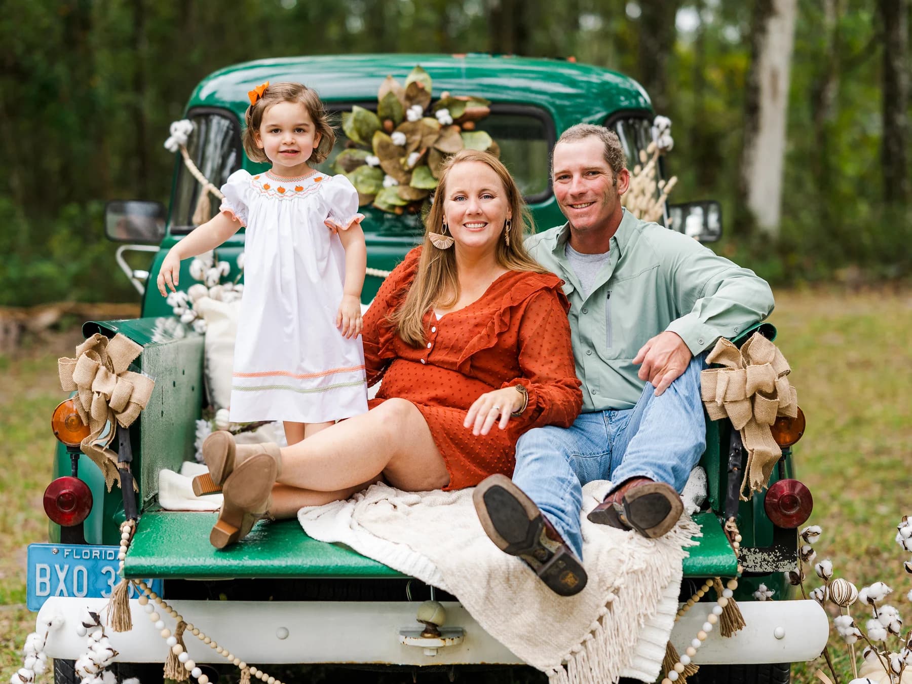 Betsy and Adam Langan sitting on the tailgate of a dark green vintage pickup with their young daughter standing beside them, the truck dressed with cotton stems, burlap bows and a magnolia wreath, pines behind.