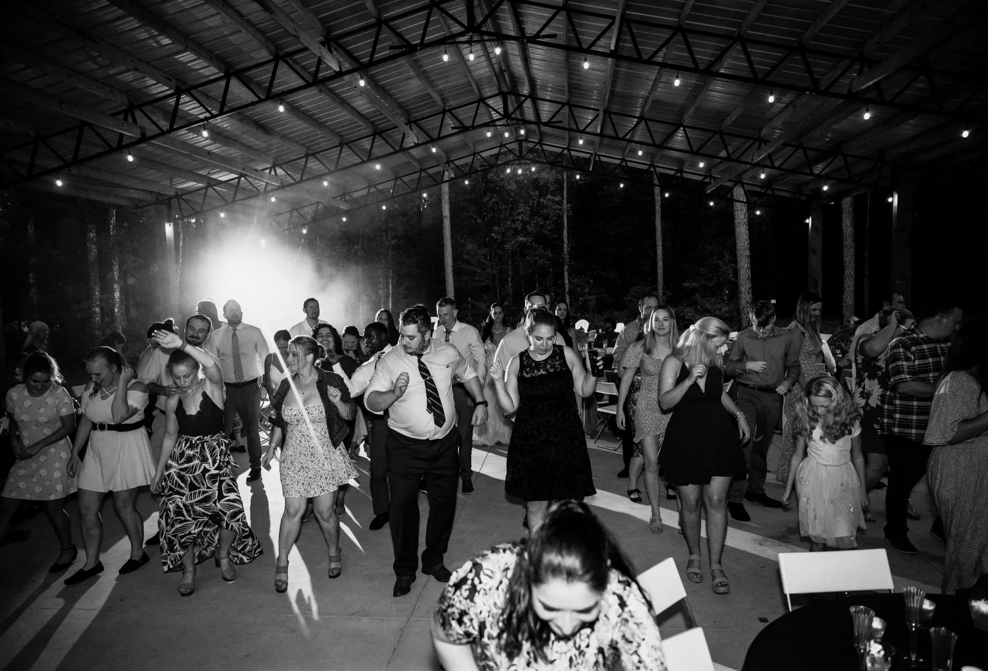 Black and white photograph of a crowded dance floor under the open sided pavilion at night, string lights and haze overhead, around twenty guests in summer dresses and shirtsleeves dancing between the steel posts.