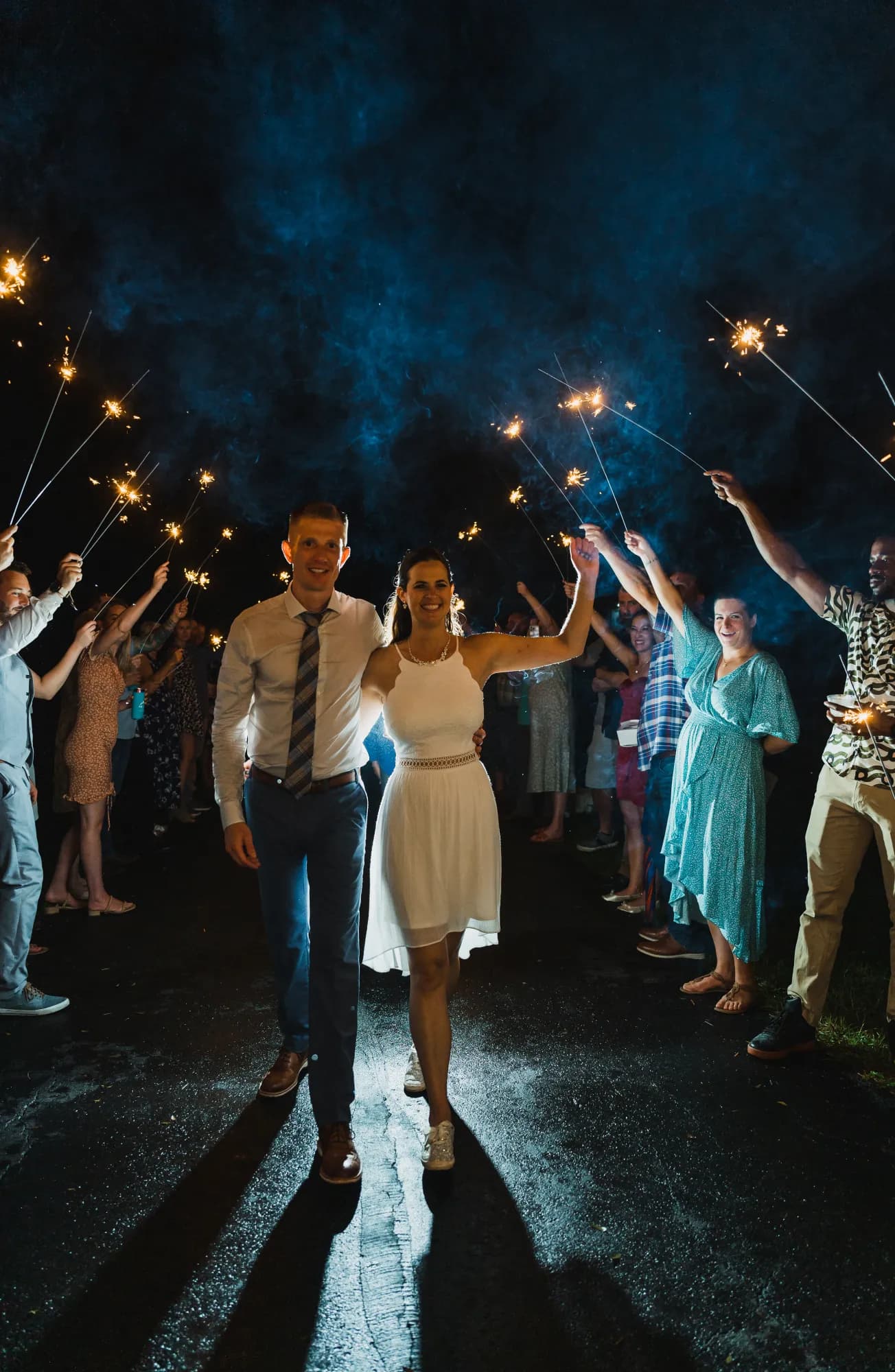A newly married couple walking hand in hand down a paved drive at night between two lines of guests holding lit sparklers overhead, blue smoke drifting through the light behind them.