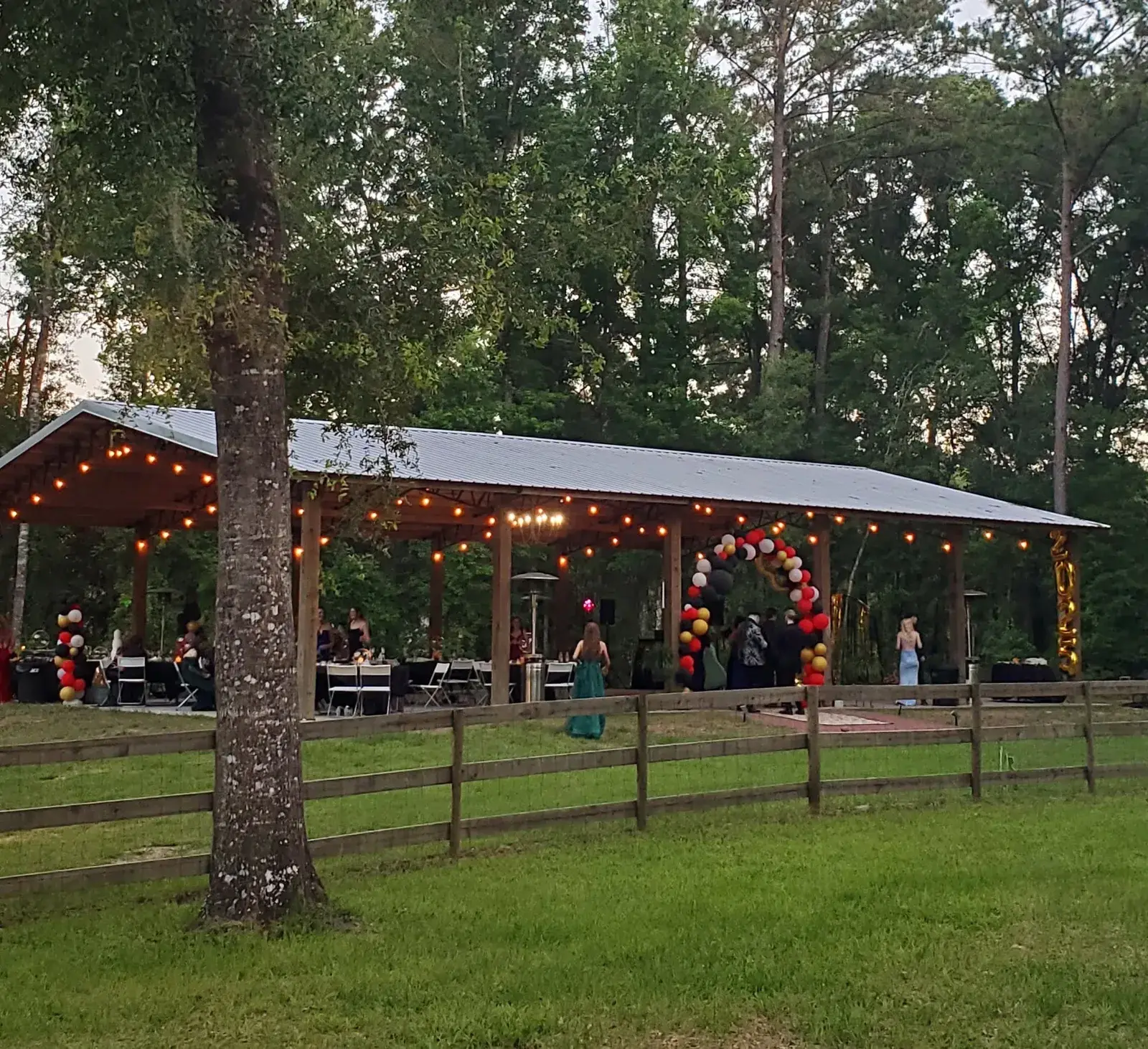 The open pavilion seen from across the pasture fence at dusk, string lights on along its whole length, a balloon arch and set tables at the far end, oaks in front.