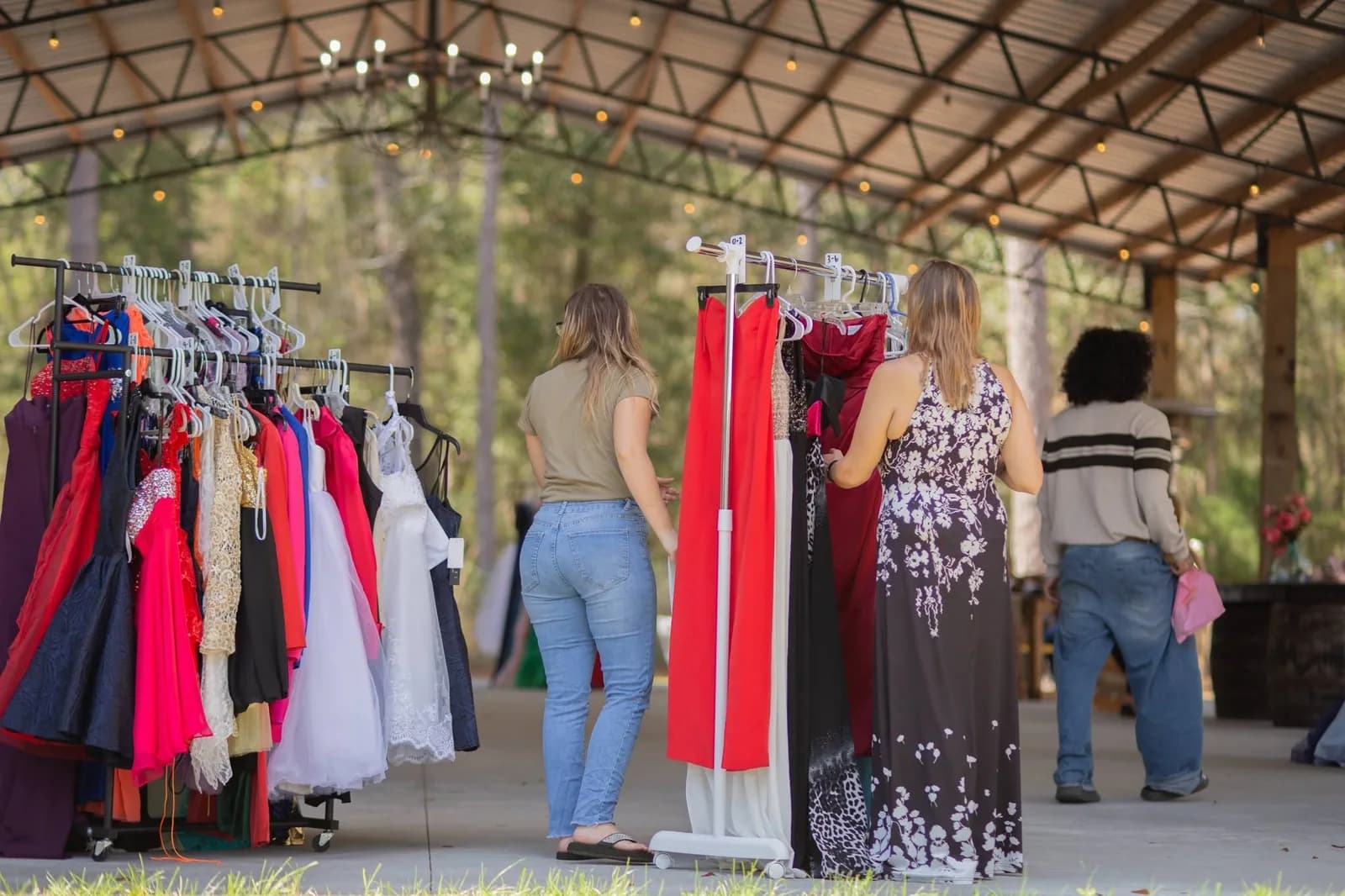 Four women looking through rails of prom dresses hung under the pavilion, the racks running along the open edge with the lawn and trees beyond.