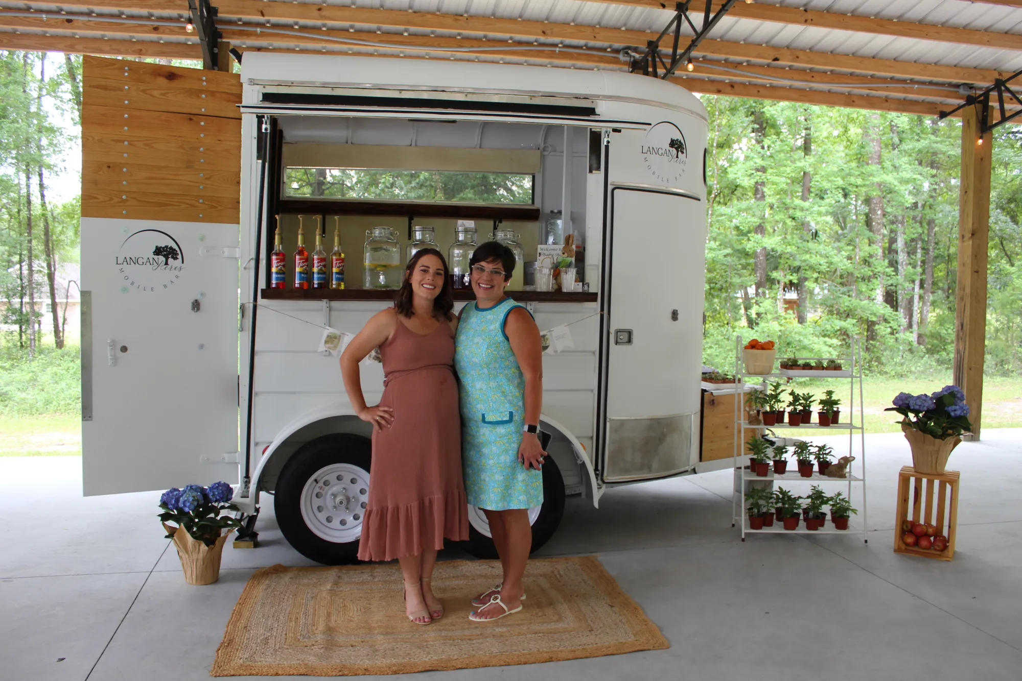 Two women standing arm in arm on a jute rug in front of the white mobile bar trailer, its hatch open to a counter of syrup bottles and glass dispensers, with tiered shelves of small potted plants and blue hydrangeas arranged on either side.