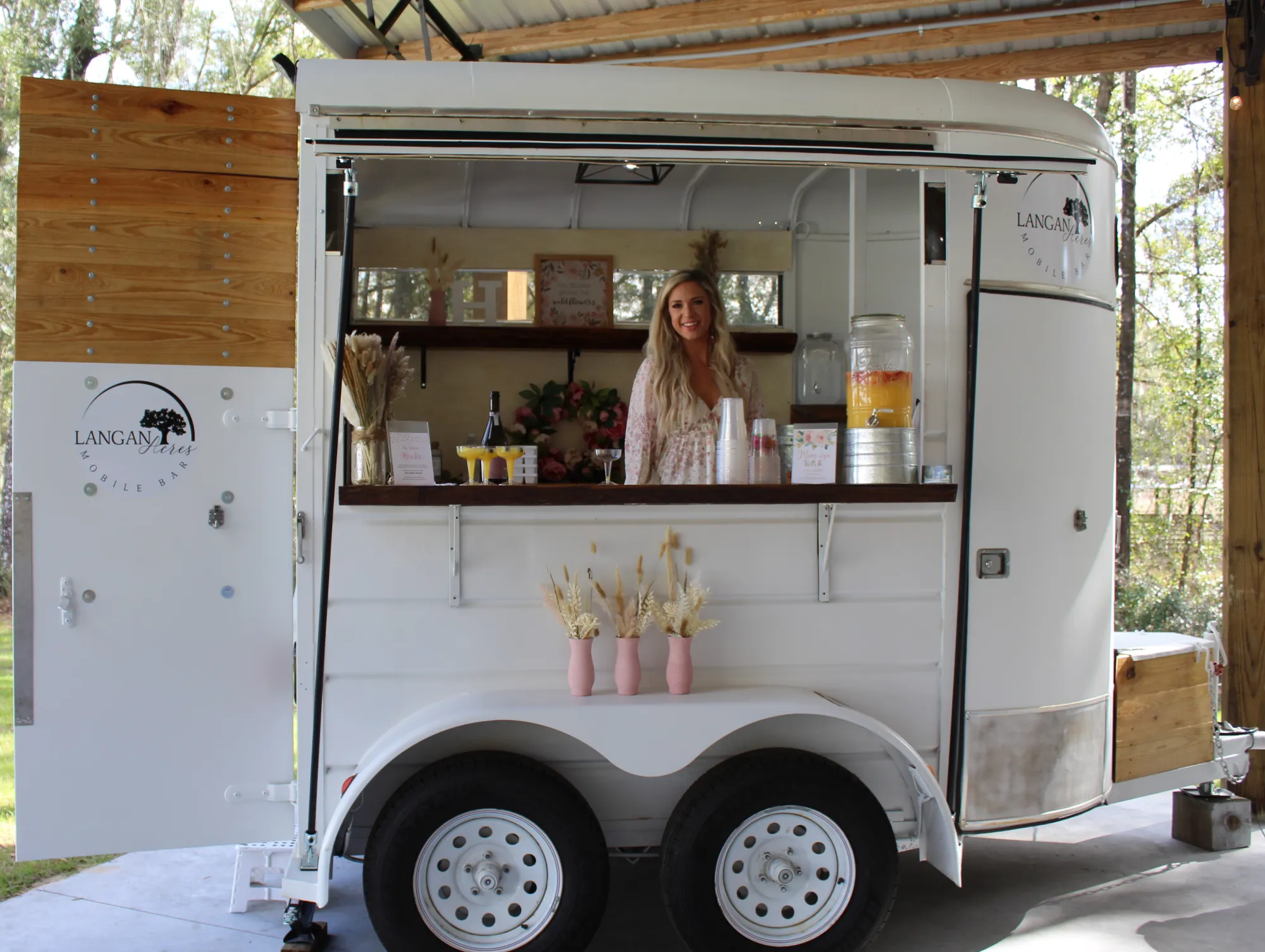A woman standing inside the white mobile bar trailer behind its wooden counter, pouring from a glass drink dispenser, the shelves behind her styled with dried grasses and a red floral arrangement and small pink vases lined along the counter front.
