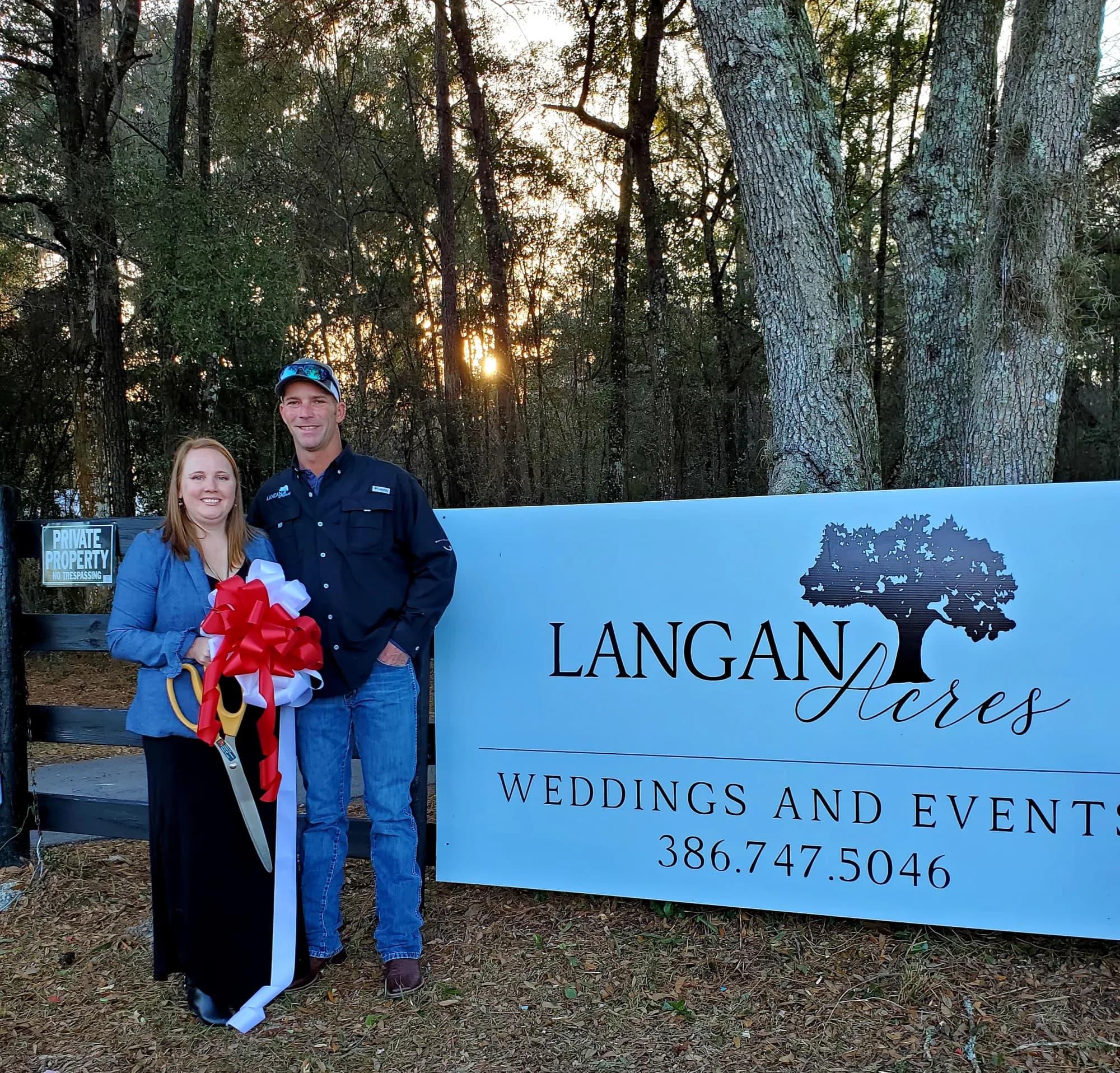 Betsy and Adam standing together beside the Langan Acres road sign at sunset, a large red bow on the sign and oversized ceremonial scissors held between them.