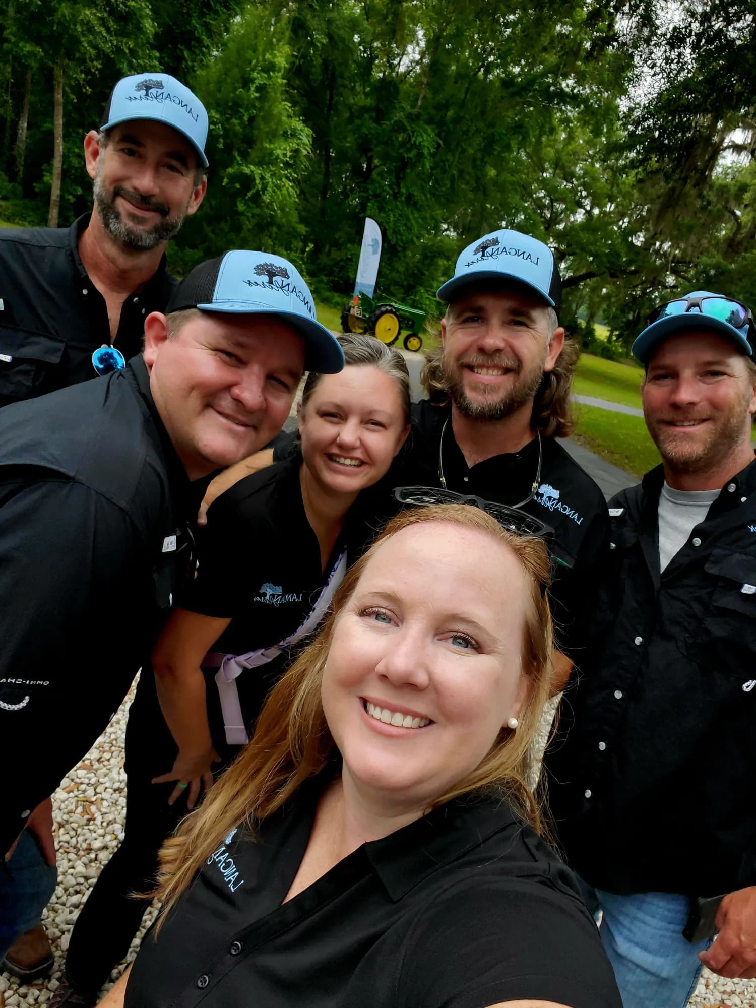 Six members of the Langan Acres team crowded into a photograph together outdoors, most in branded caps and black polo shirts, smiling at the camera.